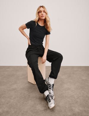 model sitting on a block wearing a black cropped t-shirt and black cargo style pants with silver sneakers against a plain background.