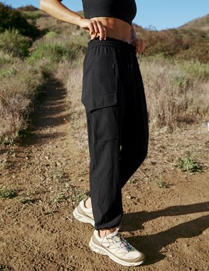 model wearing a black sports bra and black cargo style pants hiking outdoors with mountains in the background.